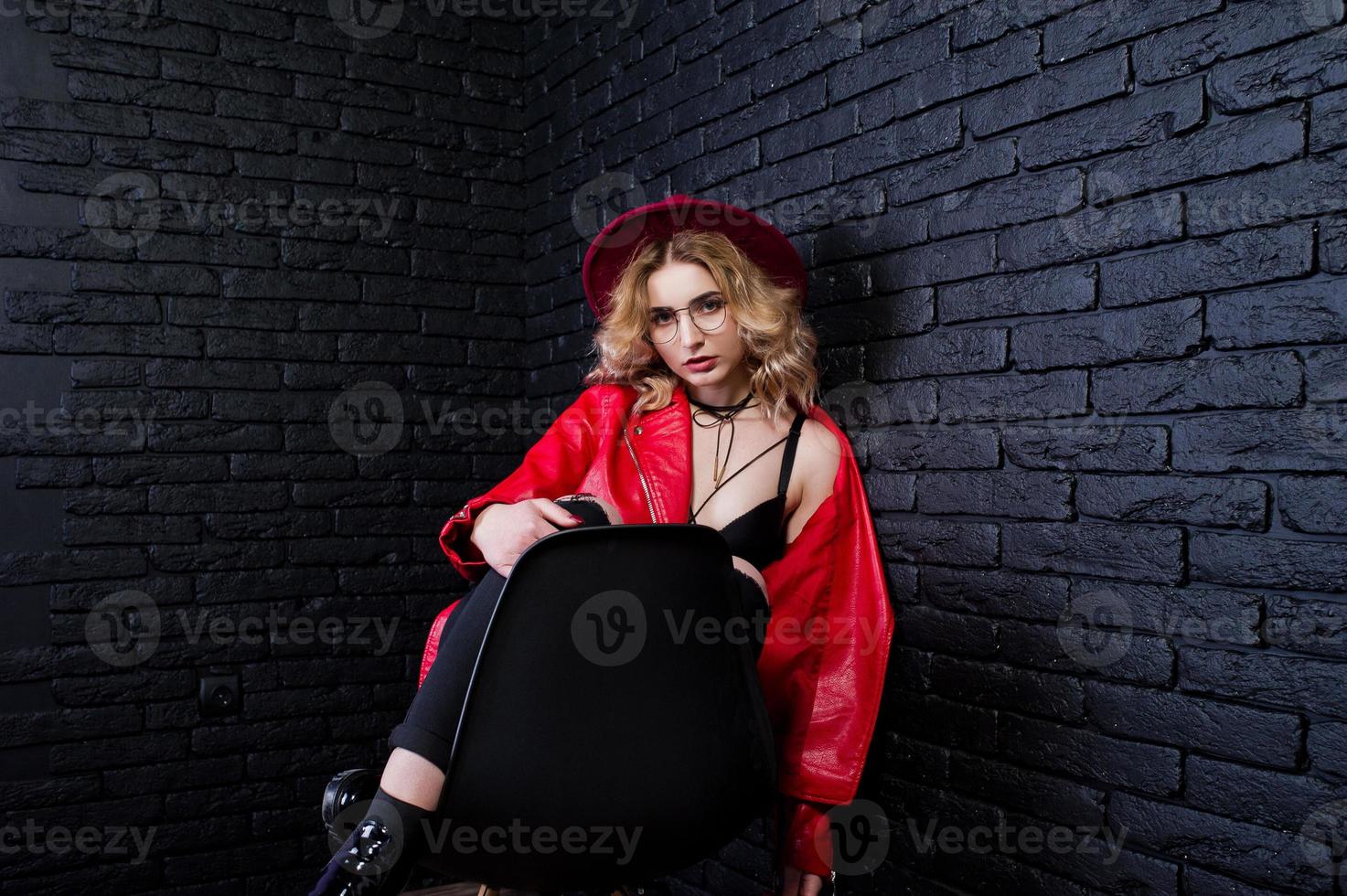 studio portret van blond meisje in rode hoed, bril en leren jas gesteld op stoel tegen bakstenen muur. foto