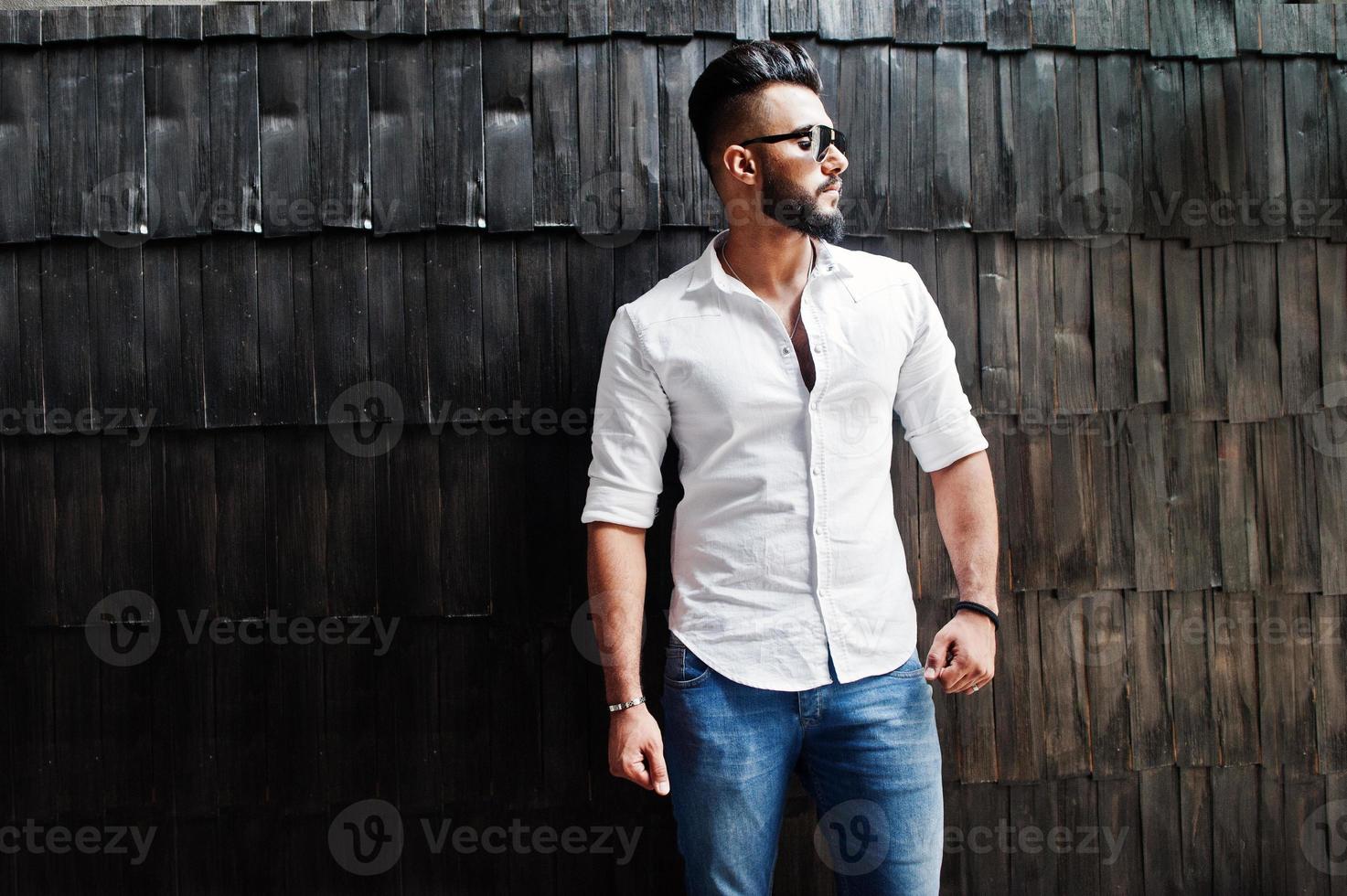 stijlvolle lange Arabische man model in wit overhemd, jeans en zonnebril poseerde tegen houten muur binnen. baard aantrekkelijke Arabische man. foto