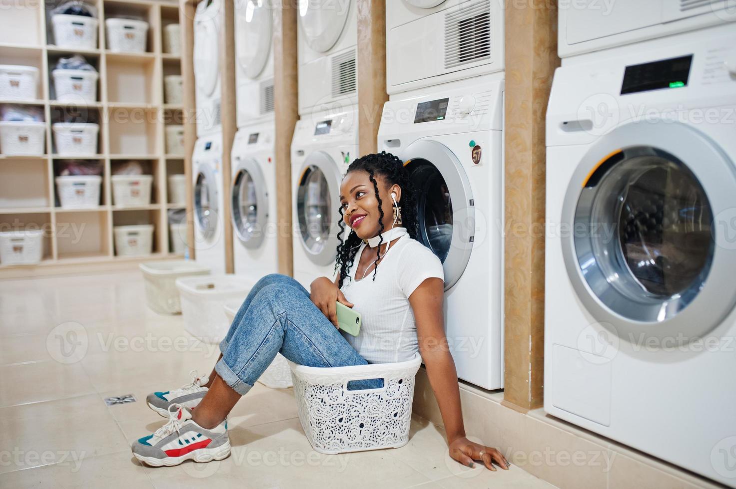 vrolijke afro-amerikaanse vrouw in de buurt van wasmachine die muziek luistert via oortelefoons van mobiele telefoon in de zelfbedieningswasserette. foto