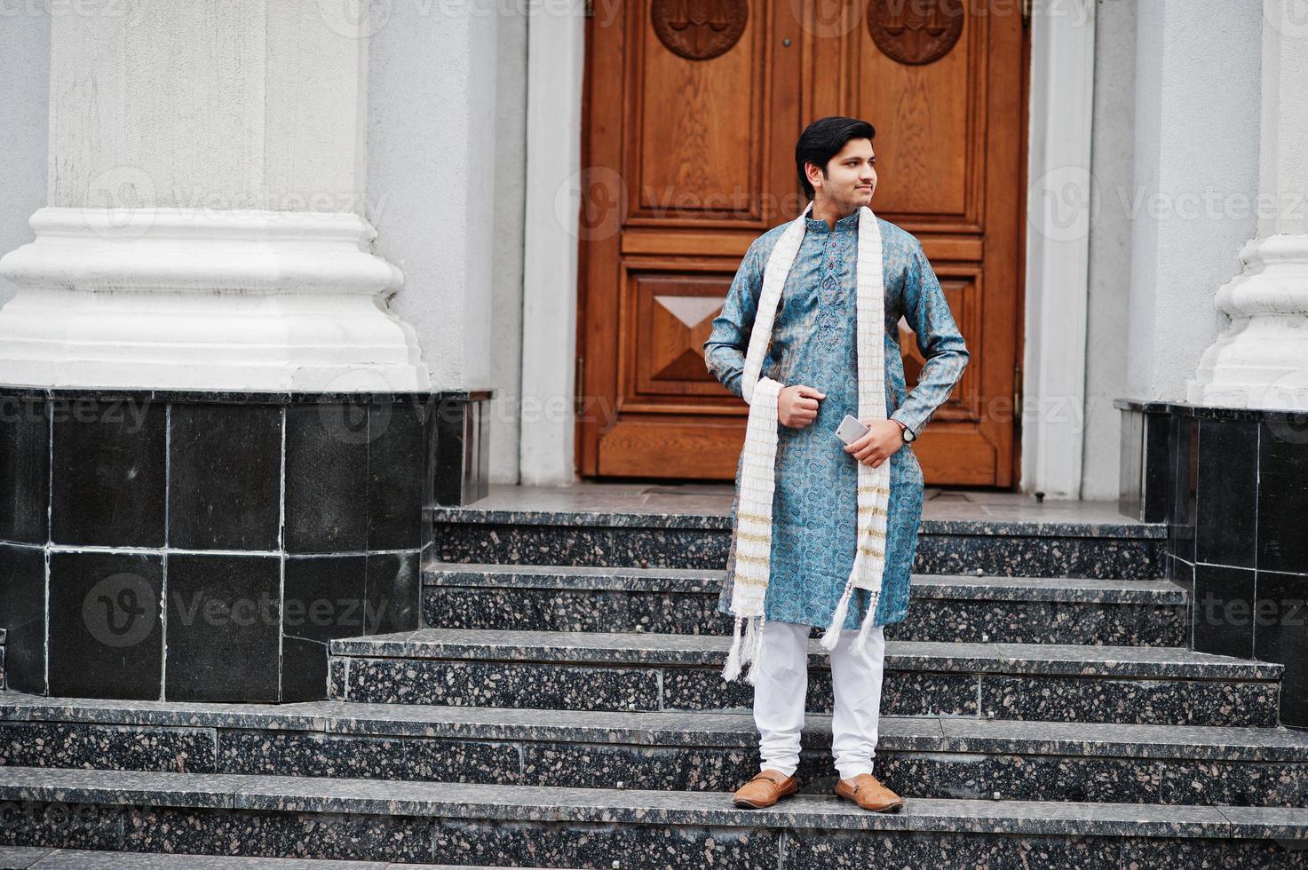 indiase man draagt traditionele kleding met witte sjaal buiten tegen de deur van het gebouw met een mobiele telefoon bij de hand. foto