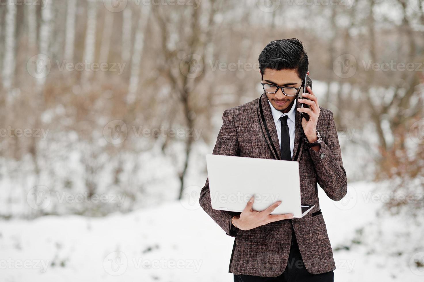 stijlvolle Indiase student man in pak en bril poseerde op winterdag buiten met laptop bij de hand, sprekend aan de telefoon. foto