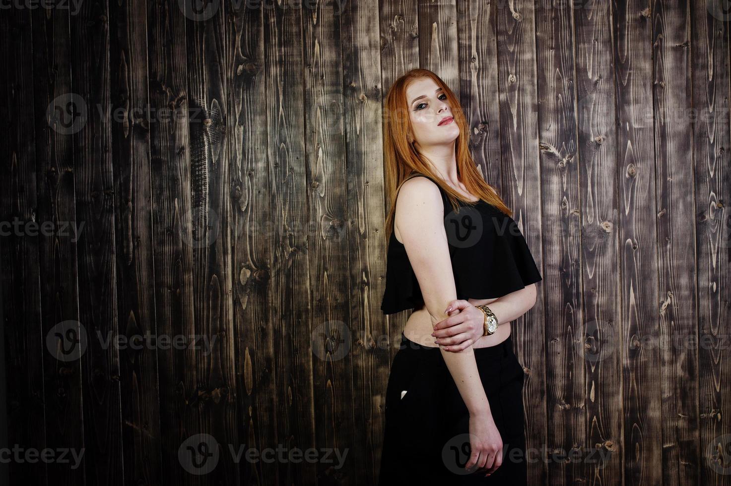 portret van een mooi roodharig meisje in zwarte top en zwarte rok poseren in de studio naast de houten muur. foto