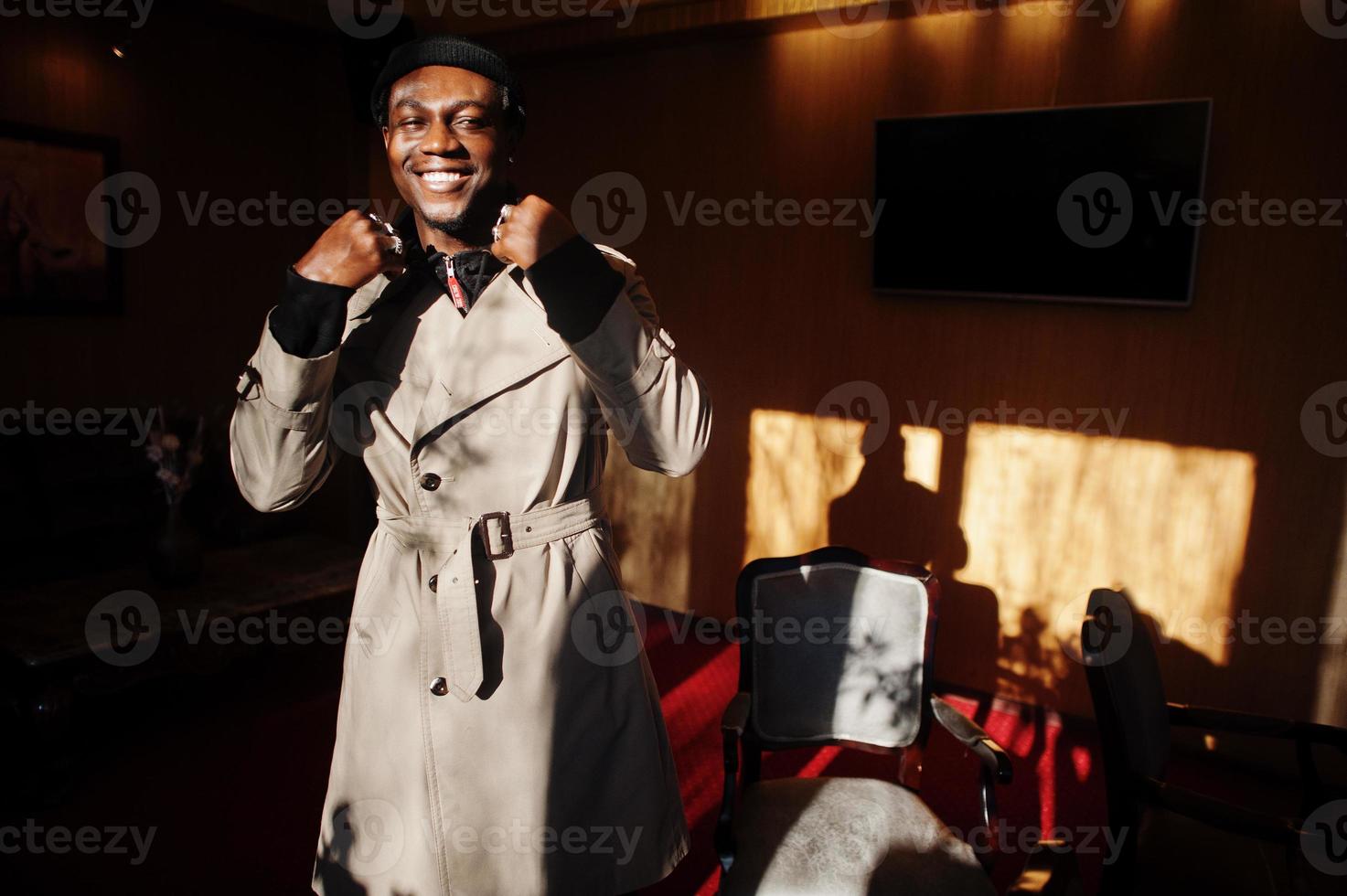 knappe Afro-Amerikaanse man poseren in kamer met zonlicht schaduwen in zwarte hoed en beige jas. foto