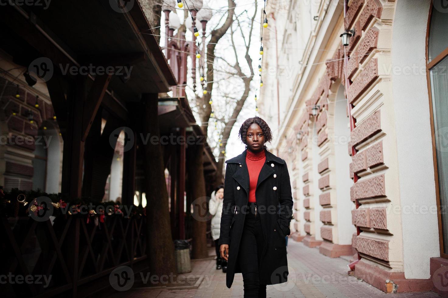 portret van een Afrikaanse vrouw met krullend haar, gekleed in een modieuze zwarte jas en rode coltrui die buiten loopt. foto