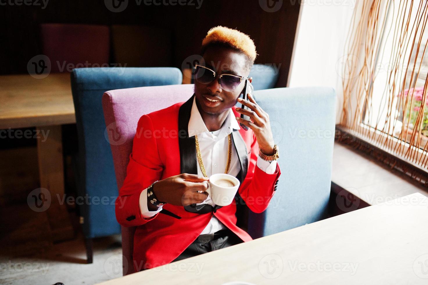 mode Afro-Amerikaanse man model in rood pak, met hoogtepunten haar en zonnebril zittend in café met koffie en spreken op mobiele telefoon. foto