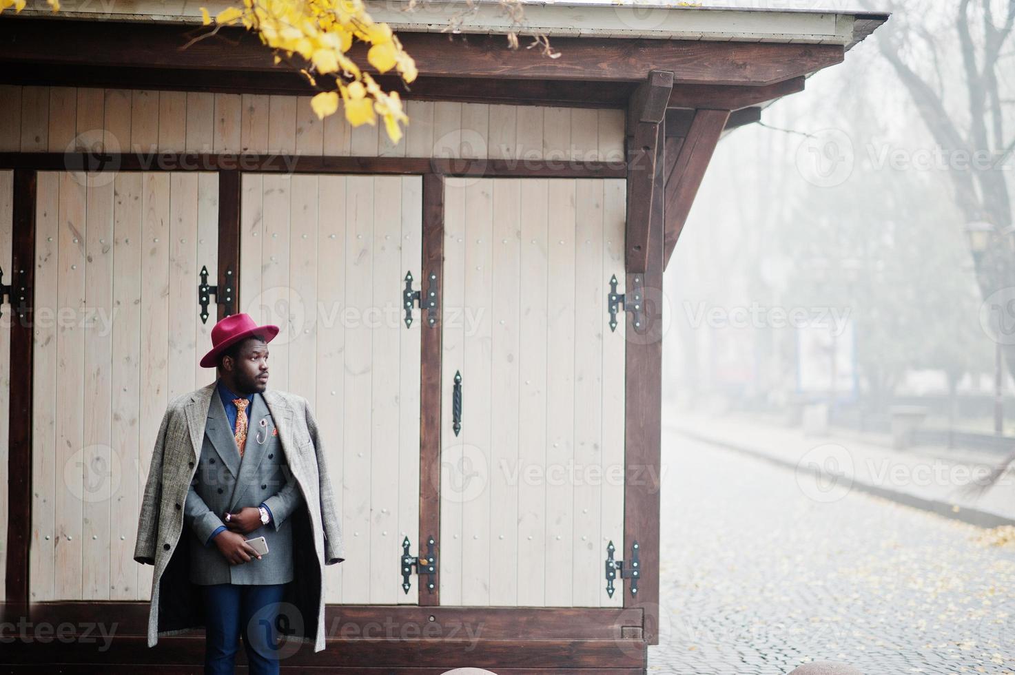 stijlvolle Afro-Amerikaanse man model in grijze jas, jas stropdas en rode hoed gesteld op mistig weer straat. foto