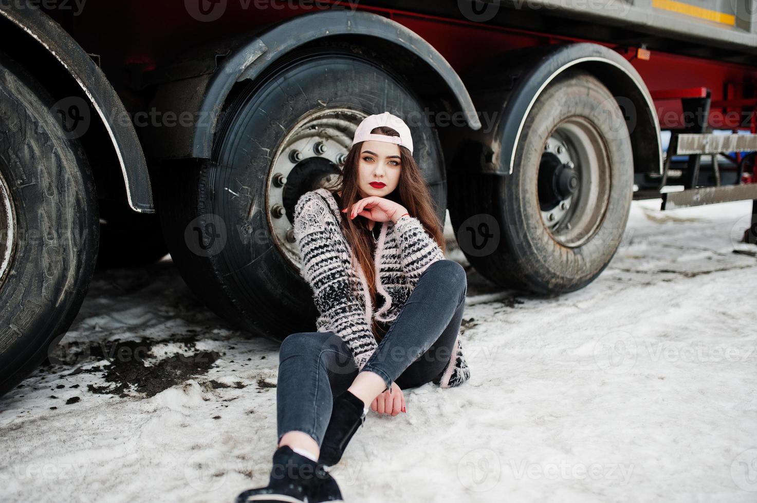 brunette stijlvolle casual meisje in cap zittend tegen vrachtwagen wielen. foto