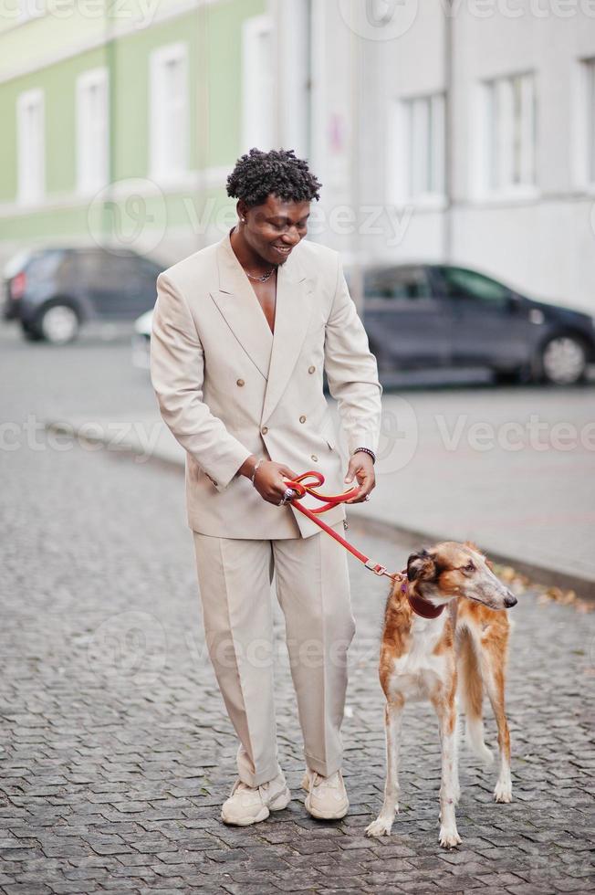 stijlvolle afro man in beige old school pak met russische borzoi hond. modieuze jonge Afrikaanse man in casual jas op blote torso. foto