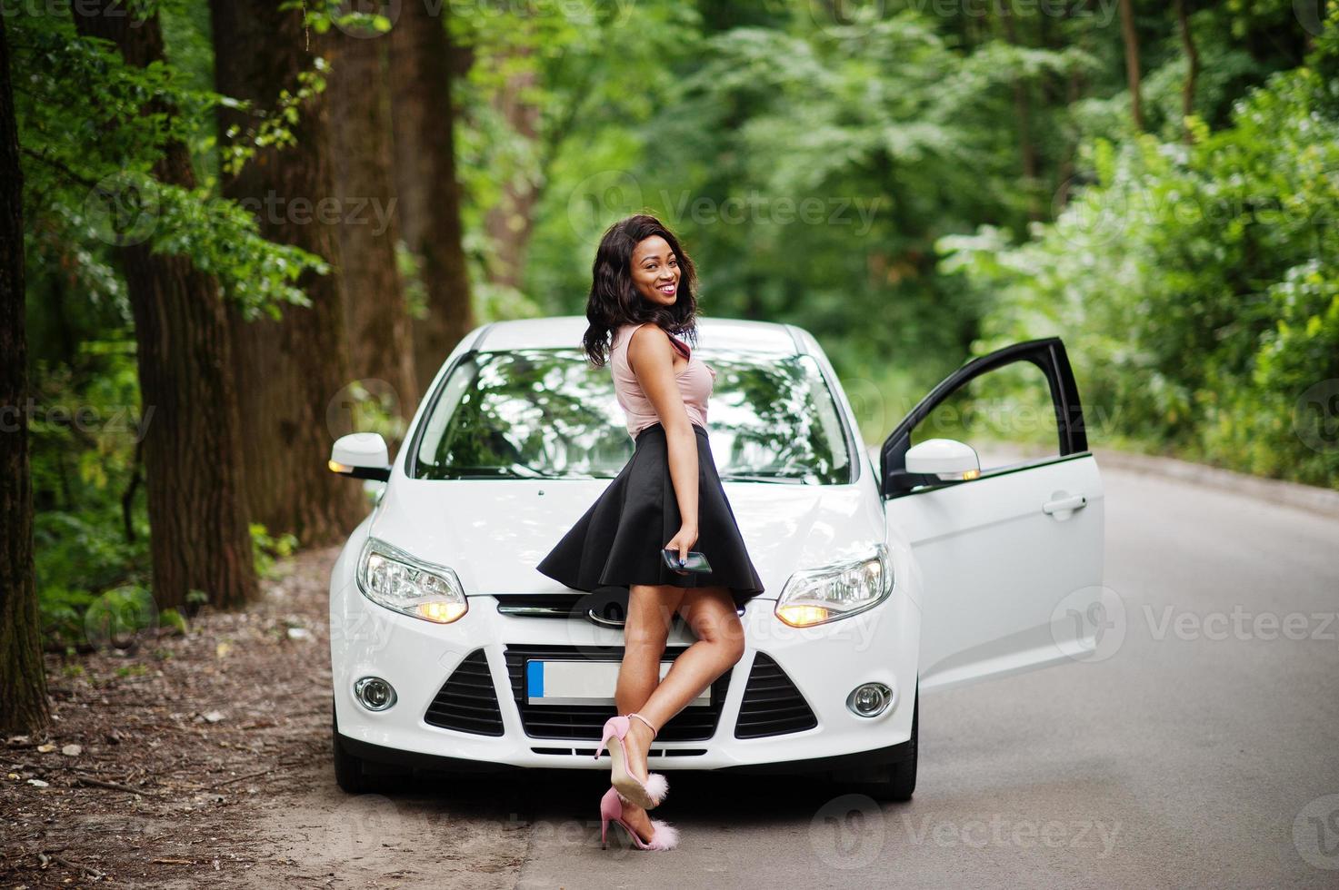 Afro-Amerikaanse vrouw poseerde tegen witte auto in bosweg. foto
