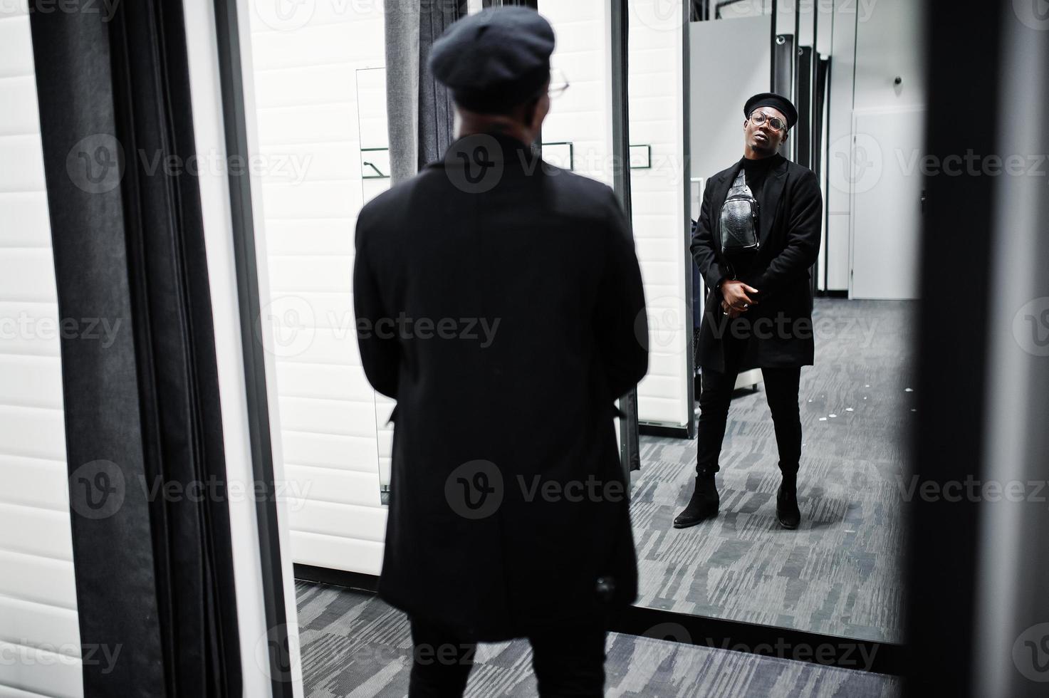 stijlvolle casual Afro-Amerikaanse man op zwarte baret en overjas met heuptas bij paskamer kledingwinkel, kijkend op spiegel. foto