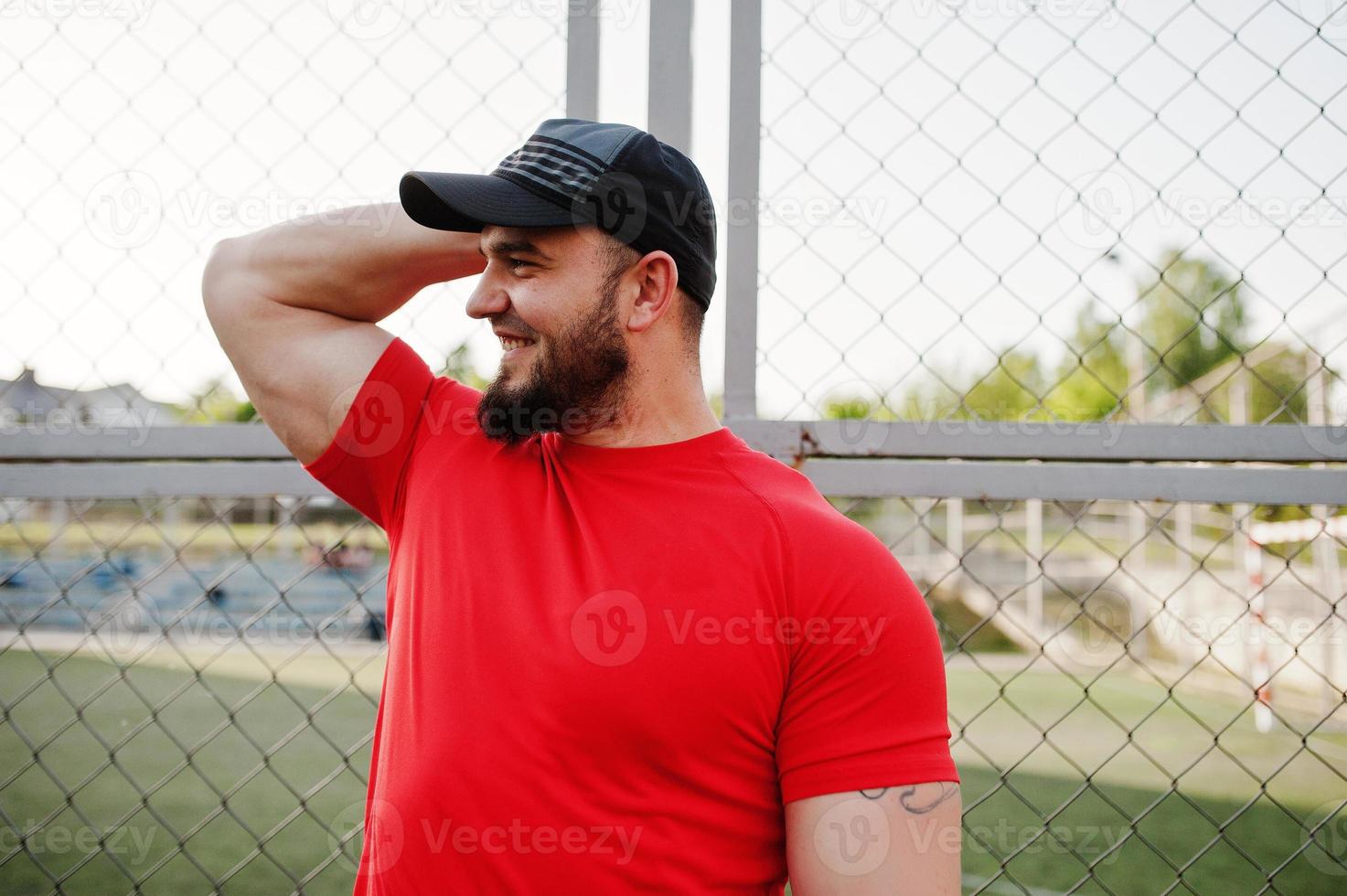 jonge brute bebaarde gespierde man draagt een rood shirt, korte broek en pet in het stadion. foto