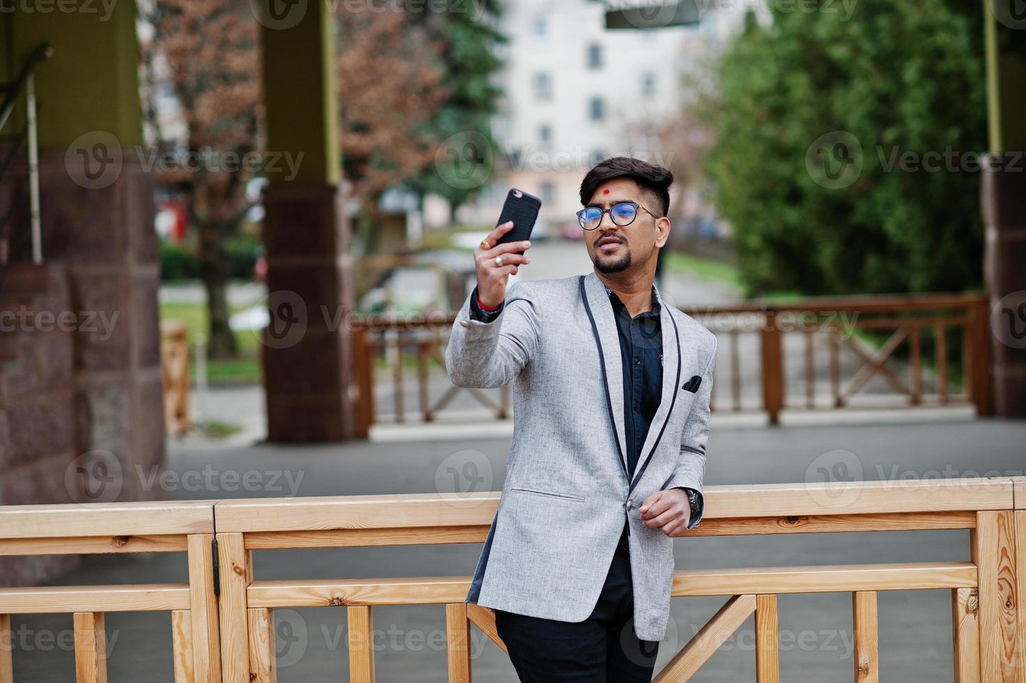 stijlvolle Indiase zakenman met bindi op voorhoofd en bril, draag op grijs pak buiten geposeerd en selfie maken op mobiele telefoon. foto