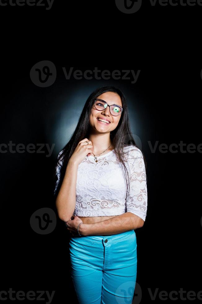 portret van een aantrekkelijke jonge vrouw in witte top en blauwe broek poseren met haar bril in het donker. foto