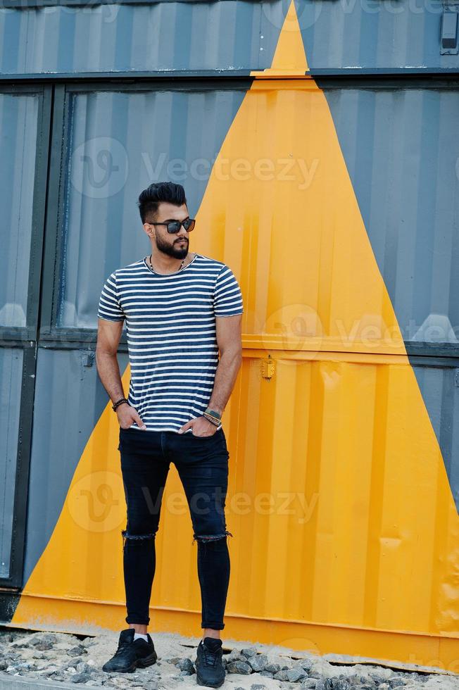 knappe lange Arabische baard man model op gestript shirt buiten geposeerd. modieuze arabische man op zonnebril tegen geel geschilderde muur. foto