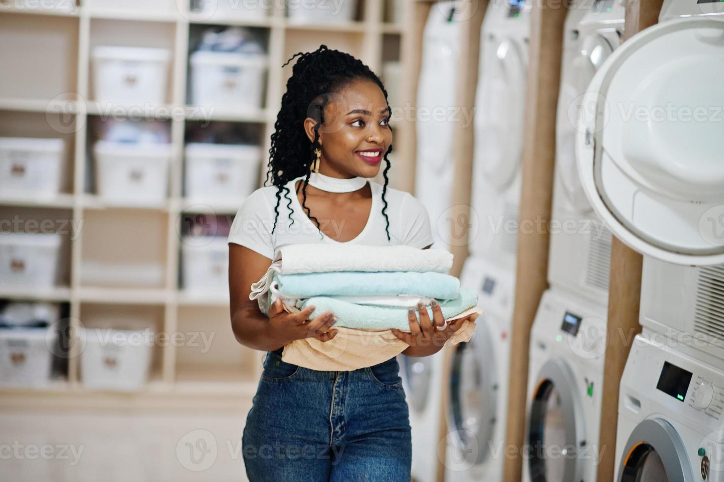 vrolijke Afro-Amerikaanse vrouw met handdoeken in handen in de buurt van wasmachine in de zelfbedieningswasserette. foto