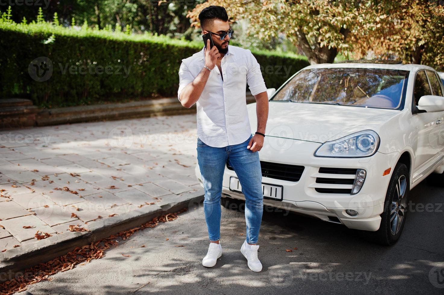 stijlvolle lange Arabische man model in wit overhemd, jeans en zonnebril gesteld op straat van de stad. baard rijke aantrekkelijke arabische man tegen witte suv auto, sprekend op mobiele telefoon. foto