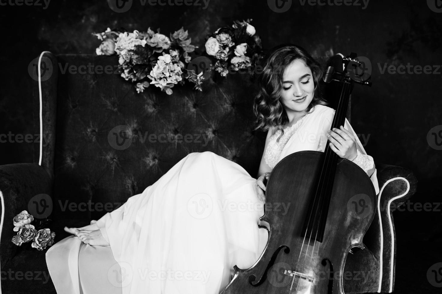 mooie jonge gilrl-muzikant in witte jurk met contrabas zittend op bruine vintage bank. foto