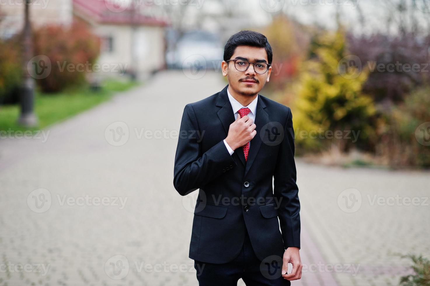 Indiase jongeman met een bril, draag een zwart pak met rode stropdas die buiten is geposeerd. foto