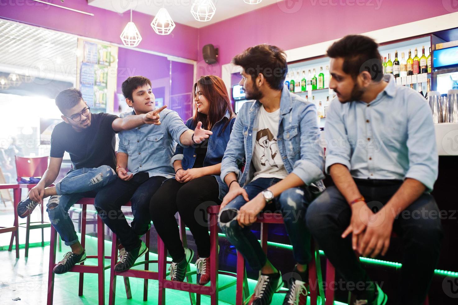 groep stijlvolle aziatische vrienden dragen jeans op stoelen tegen de bar in de club en samen chatten. foto