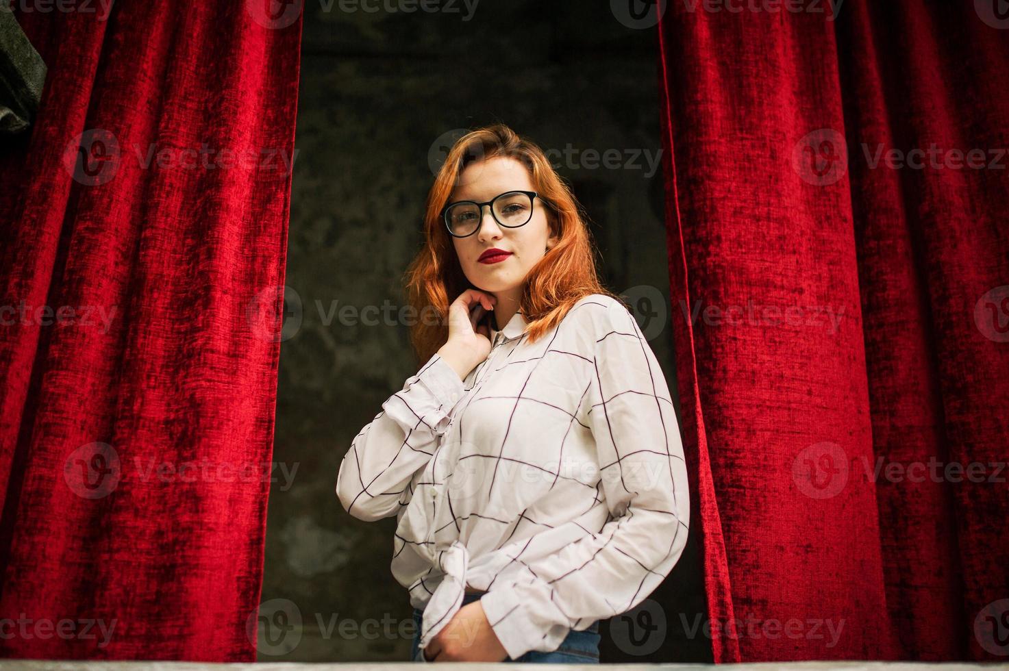 aantrekkelijke roodharige vrouw in brillen, draag op witte blouse poseren bij boog van open rode gordijnen. foto