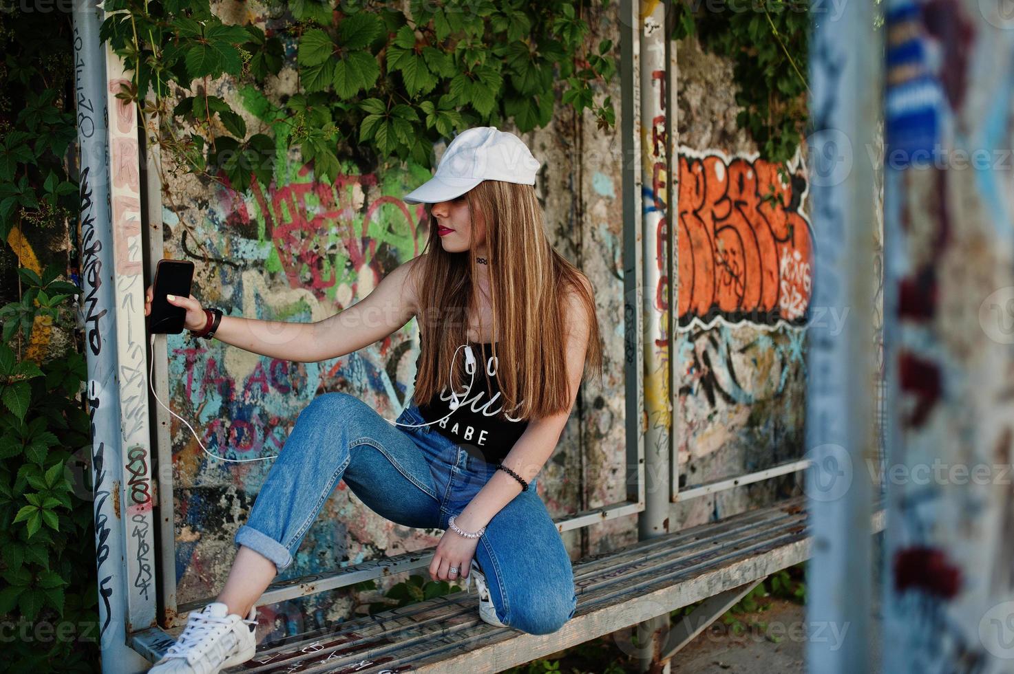 stijlvolle casual hipster meisje in pet en spijkerbroek dragen, muziek luisteren van koptelefoon van mobiele telefoon tegen grote graffiti muur. foto