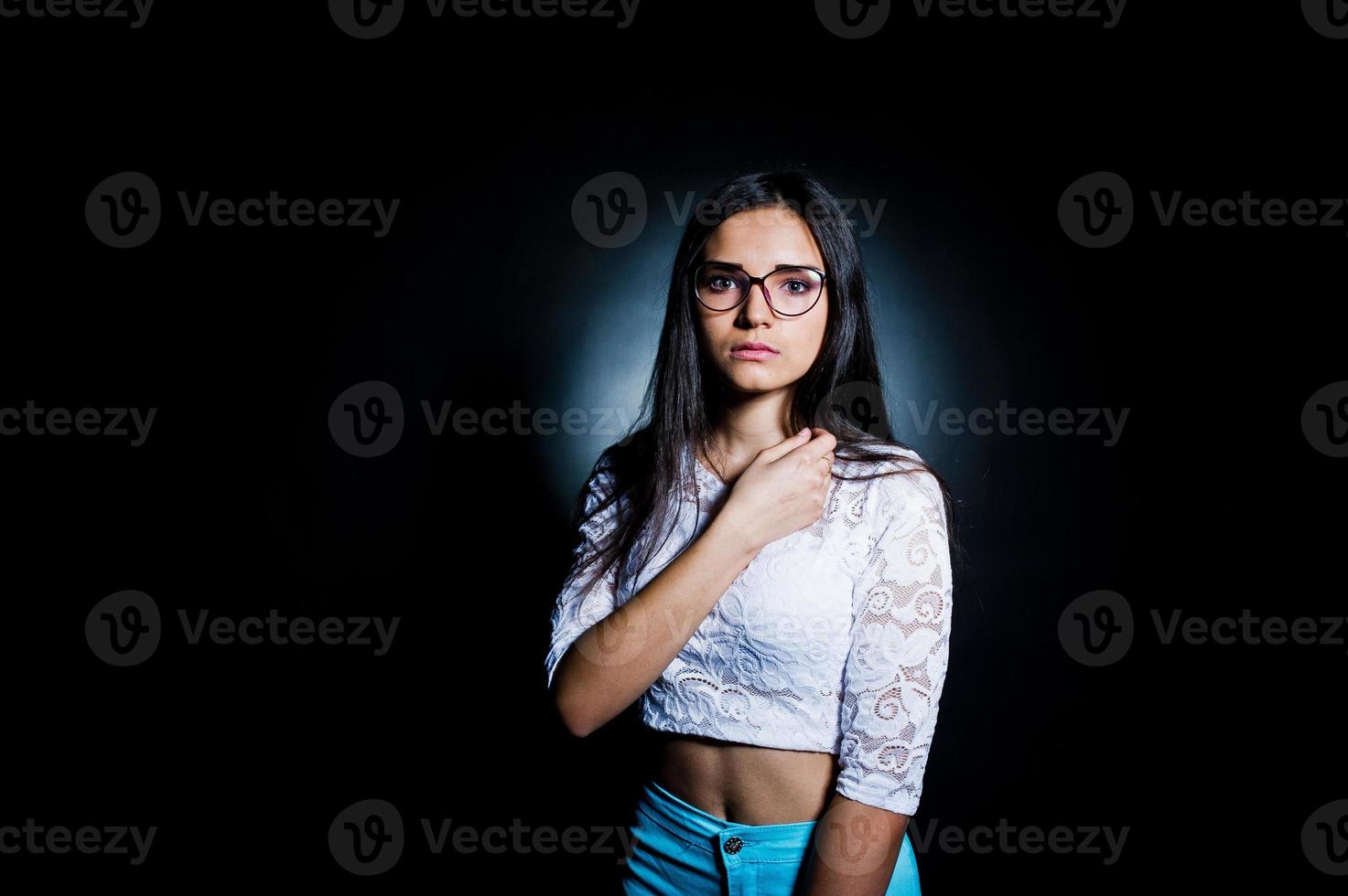 portret van een aantrekkelijke jonge vrouw in witte top en blauwe broek poseren met haar bril in het donker. foto