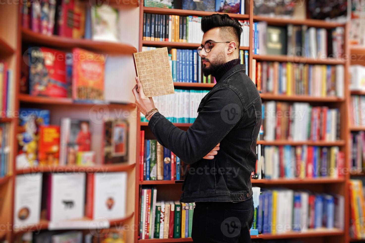 lange slimme arabische studentenman, draag een zwart spijkerjack en een bril, in de bibliotheek met een boek bij de hand. foto