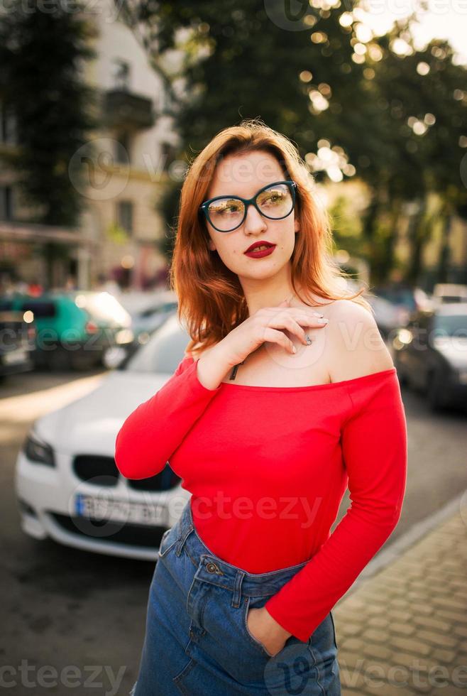aantrekkelijke roodharige vrouw in brillen, dragen op rode blouse en jeans rok poseren op straat tegen witte sportwagen. foto