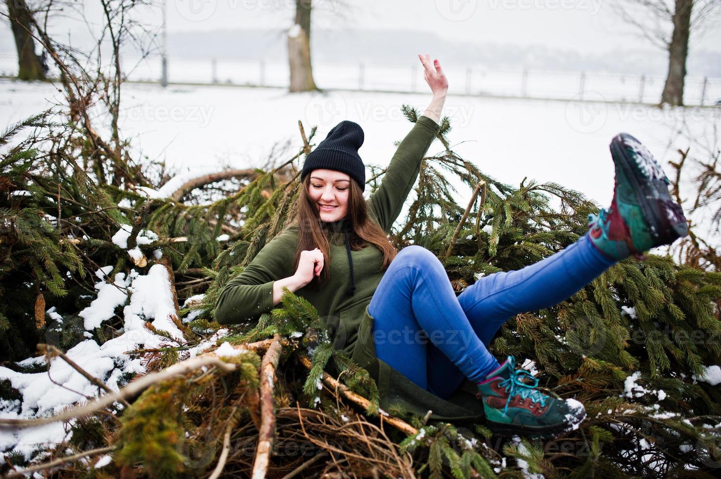 jong meisje draagt een lang groen sweatshirt, jeans en zwarte hoofddeksels aan takken van de dennenboom in de winterdag. foto