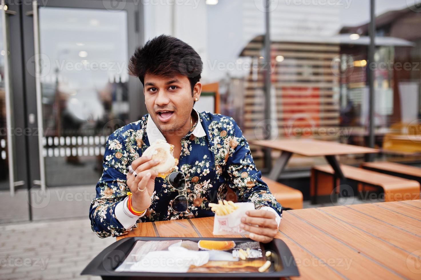 aantrekkelijke en knappe stijlvolle Indiase man draagt een shirt en eet hamburger en frietjes op het terras van het moderne loungecafé tijdens zijn werkpauze. foto