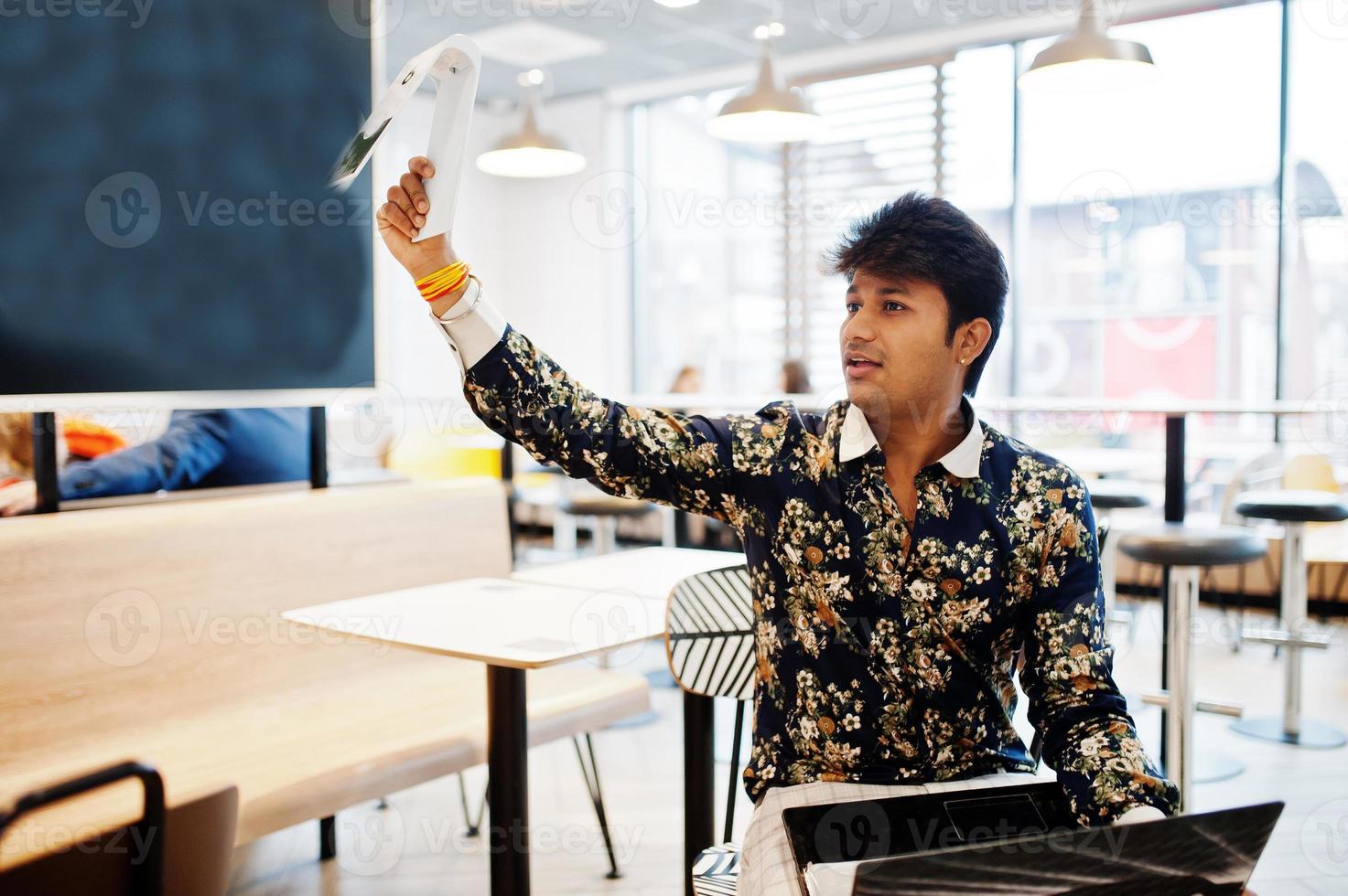 jonge mannelijke Indiase freelancer zit met opengeklapte laptopcomputer in fastfoodcafé, knappe aziatische man houdt het bord met de bestelling vast en wacht op de ober in een comfortabele coffeeshop. foto
