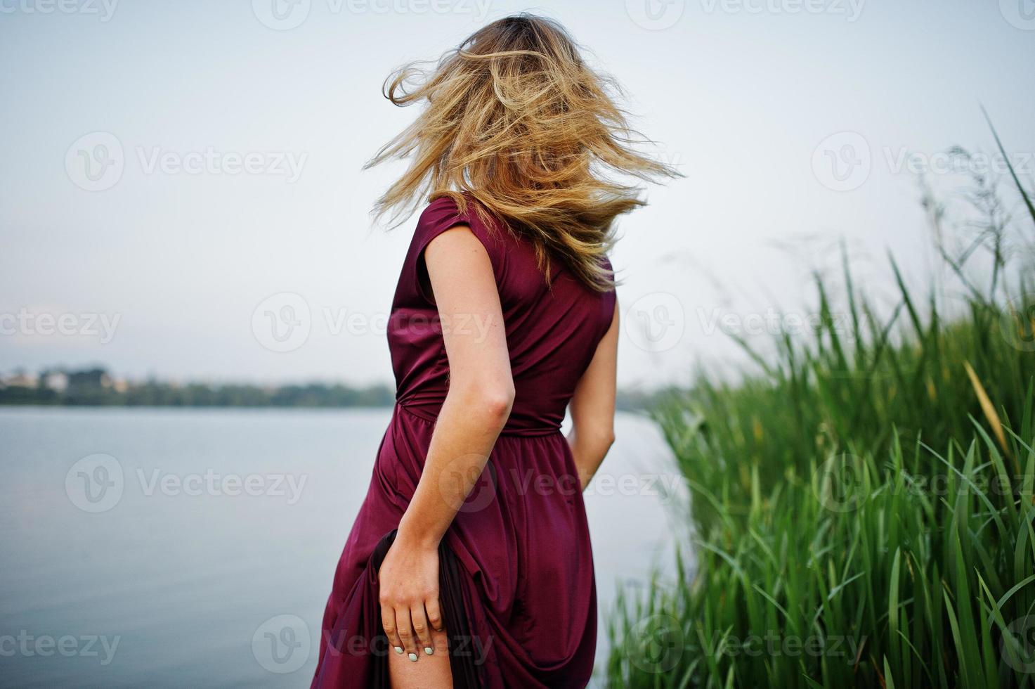 blonde sensuele vrouw in rode marsala-jurk die in het water van het meer met riet staat en haar naakte been laat zien. foto