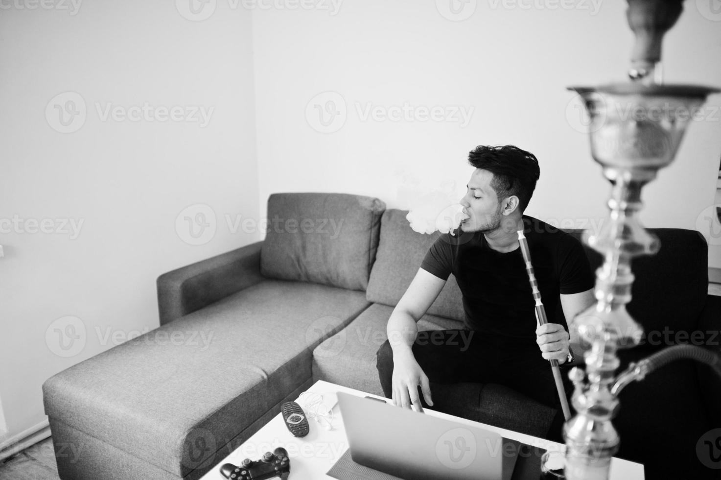 knappe en modieuze Indiase man in het zwart zittend op kamer, rokende waterpijp, werken op laptop. foto
