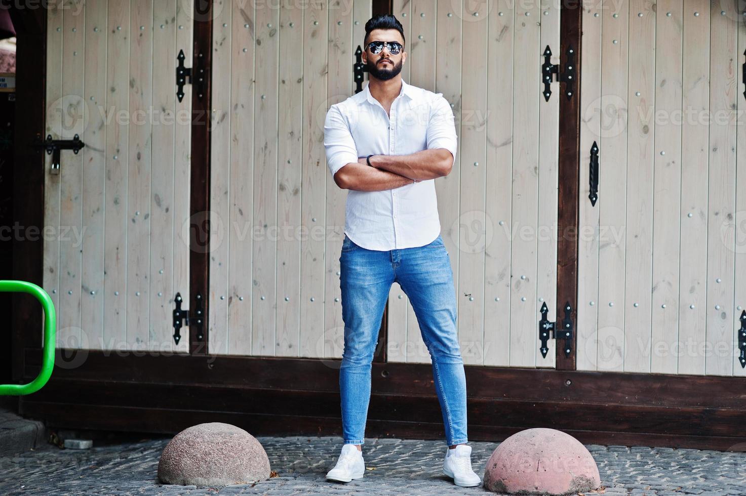 stijlvolle lange Arabische man model in wit overhemd, jeans en zonnebril gesteld op straat van de stad. baard aantrekkelijke Arabische man. foto