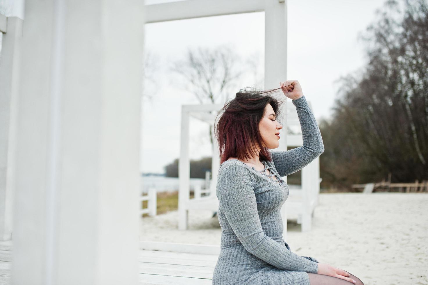 portret van brunette meisje in grijze jurk zit op witte houten constructie. foto