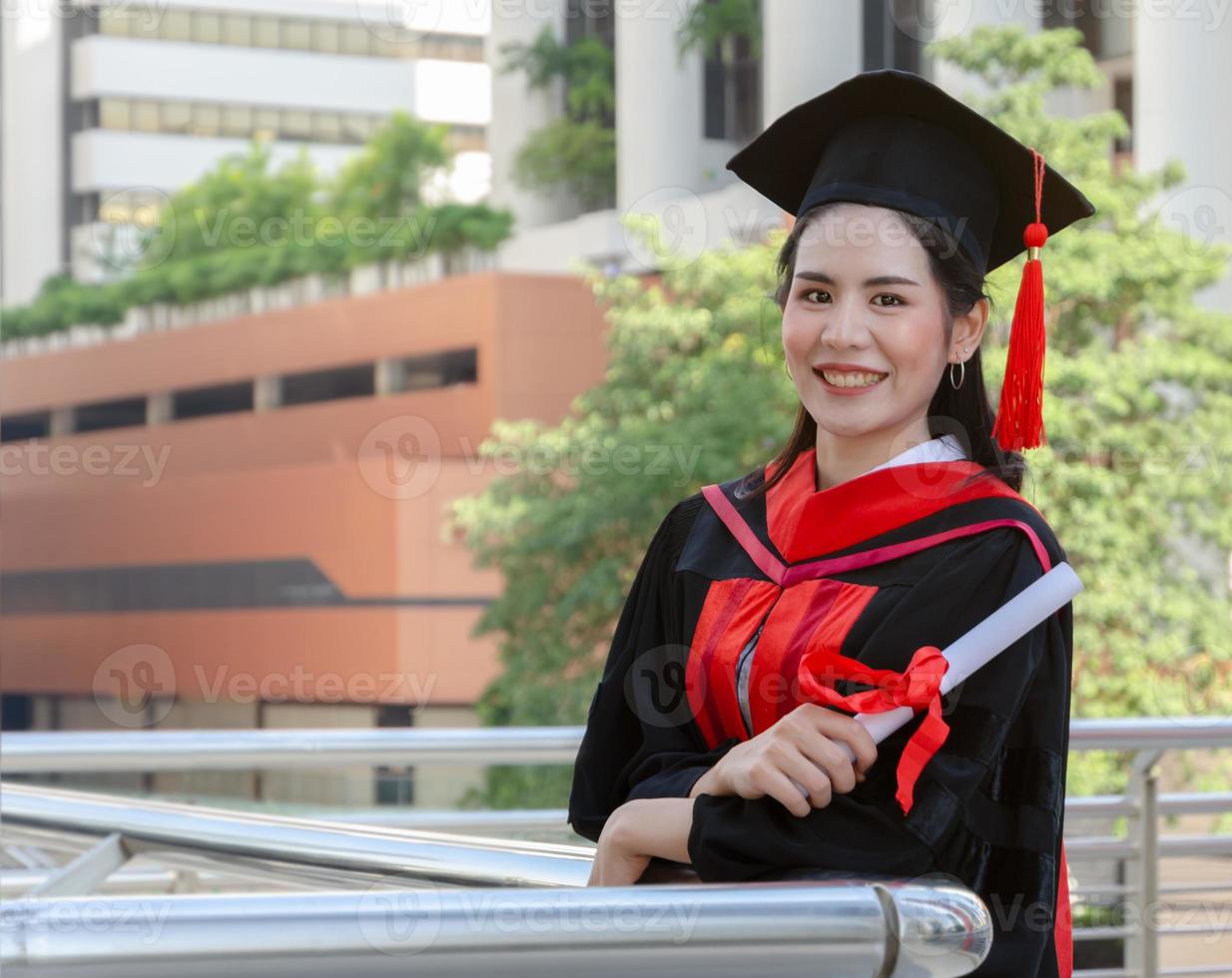 gediplomeerde vrouw die baret en academische kleding draagt. glimlachende student die zich met gekruiste wapens bevindt en diploma houdt foto