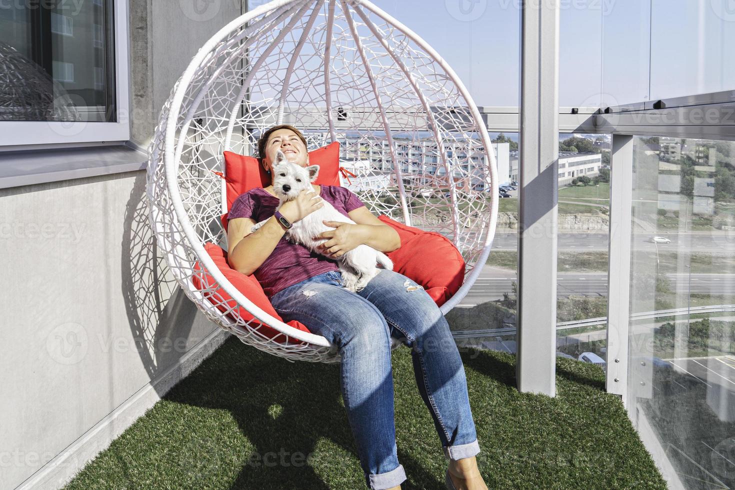 duizendjarige vrouw met witte puppy zittend in moderne hangende stoel op balkon van flatgebouw en glimlach. foto