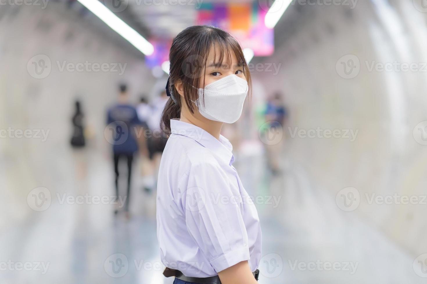 mooie jonge aziatische student in uniform loopt in een ondergrondse tunnel om 's ochtends de metro naar school te nemen terwijl hij een masker draagt voor stof, covid-19-bescherming en goede hygiëne. foto
