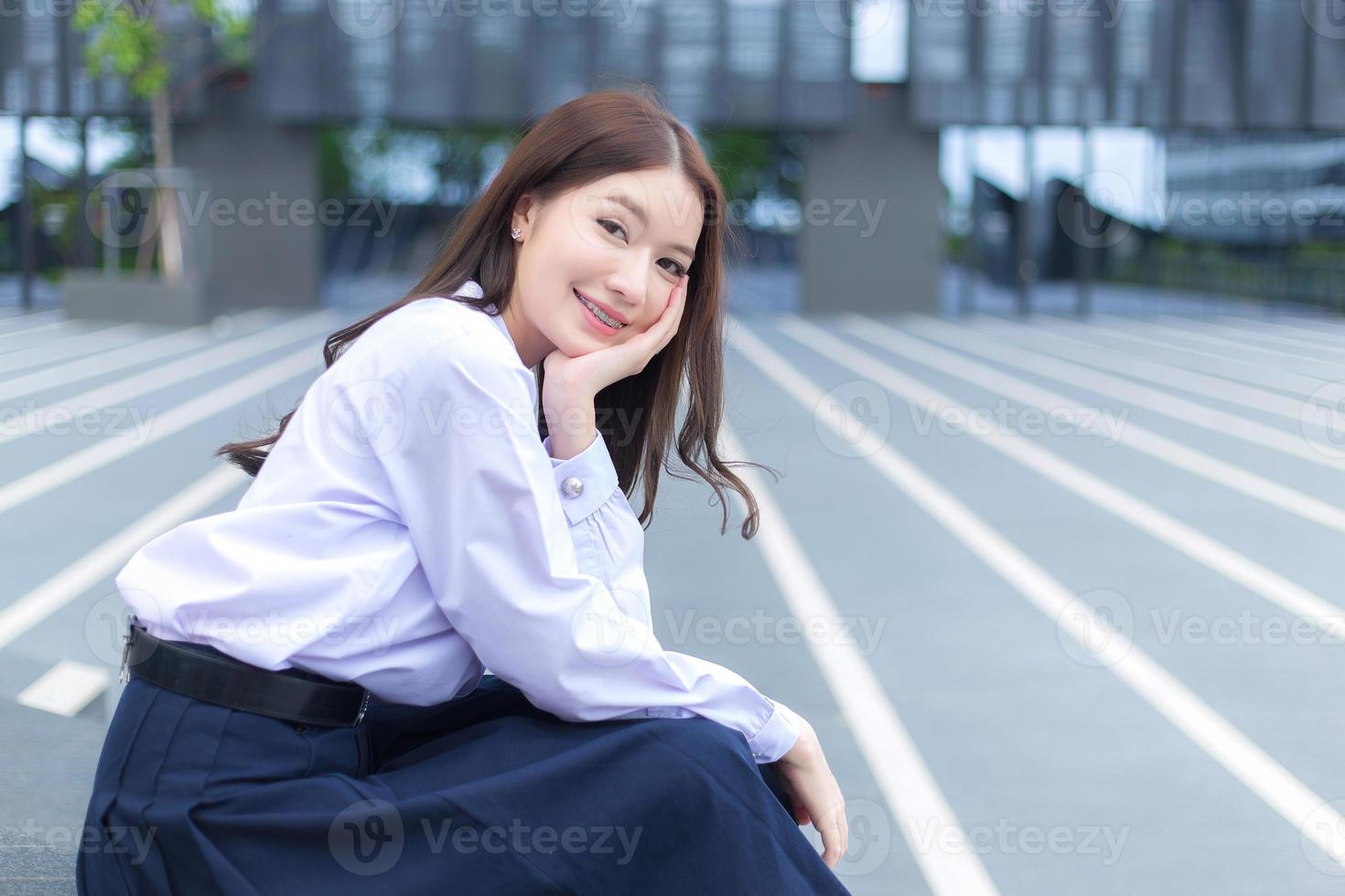mooie Aziatische middelbare school student meisje in het schooluniform met beugels op haar tanden zit en glimlacht vol vertrouwen terwijl ze gelukkig naar de camera kijkt met het gebouw op de achtergrond. foto