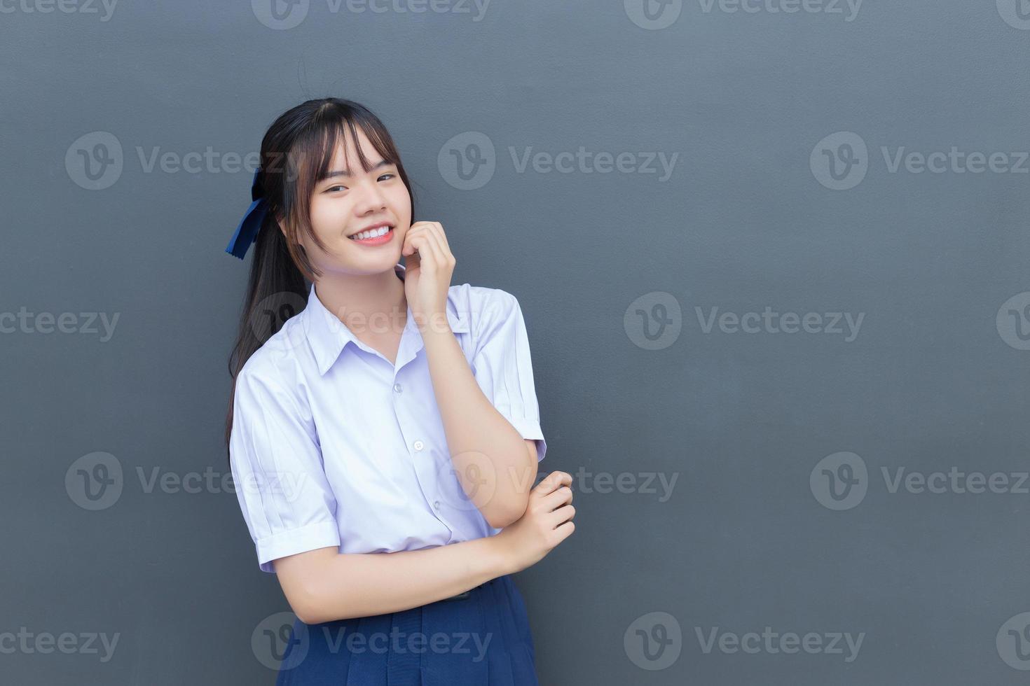 mooie Aziatische middelbare school student meisje in het schooluniform met een zelfverzekerde glimlach terwijl ze gelukkig naar de camera kijkt met grijs op de achtergrond. foto