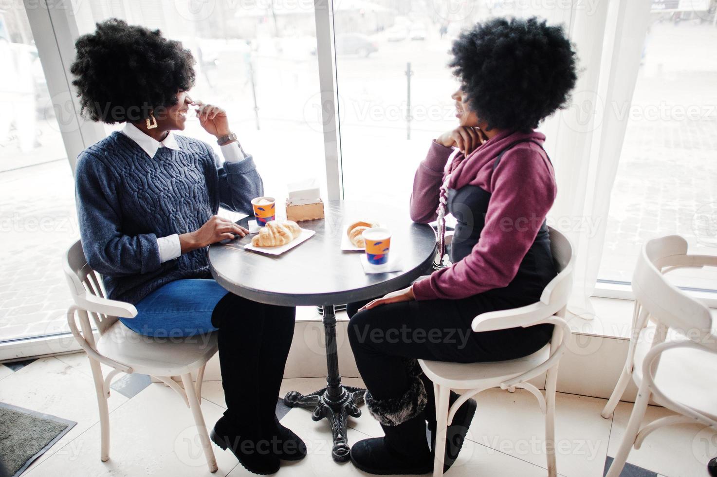 twee Afro-Amerikaanse vrouwen met krullend haar dragen op truien zit aan tafelcafé, eet een croissant en drinkt thee. foto