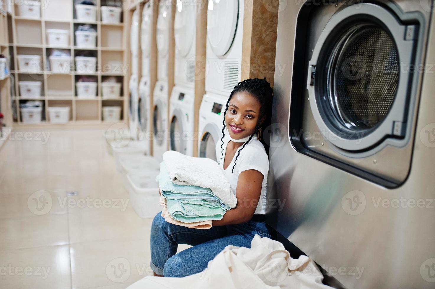 vrolijke Afro-Amerikaanse vrouw met handdoeken in handen in de buurt van wasmachine in de zelfbedieningswasserette. foto