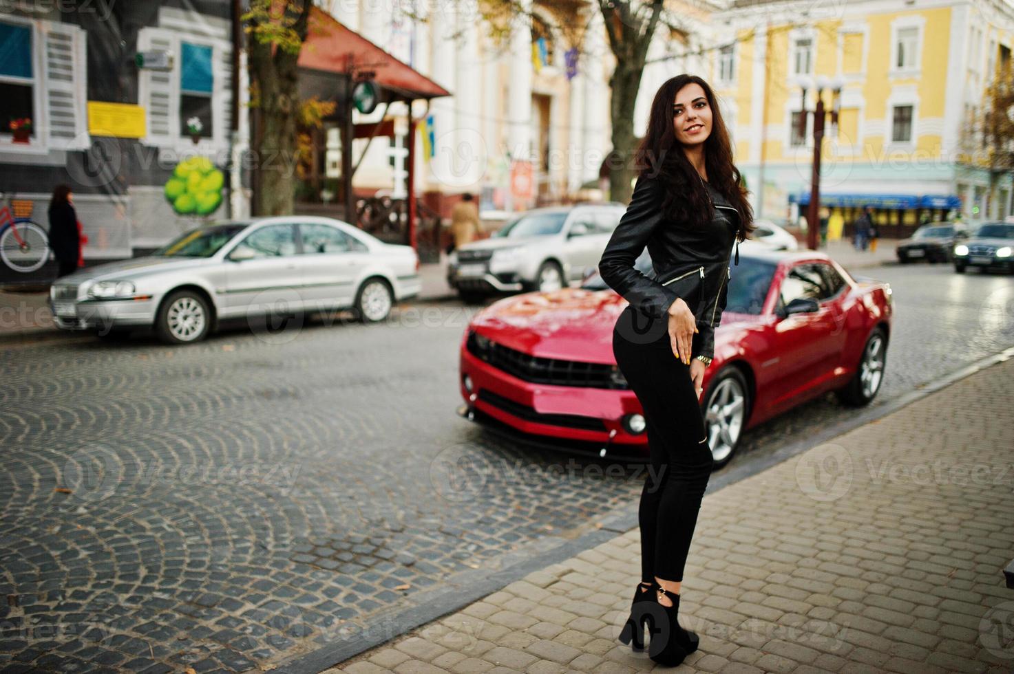 jonge krullend en sexy vrouw in leren jas tegen rode muscle car op straat. foto