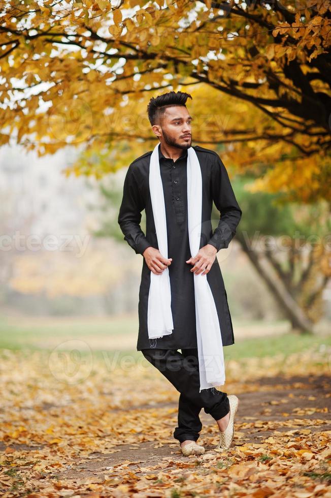 Indiase stijlvolle man in zwarte traditionele kleding met witte sjaal buiten poseerde tegen gele herfstbladeren boom. foto