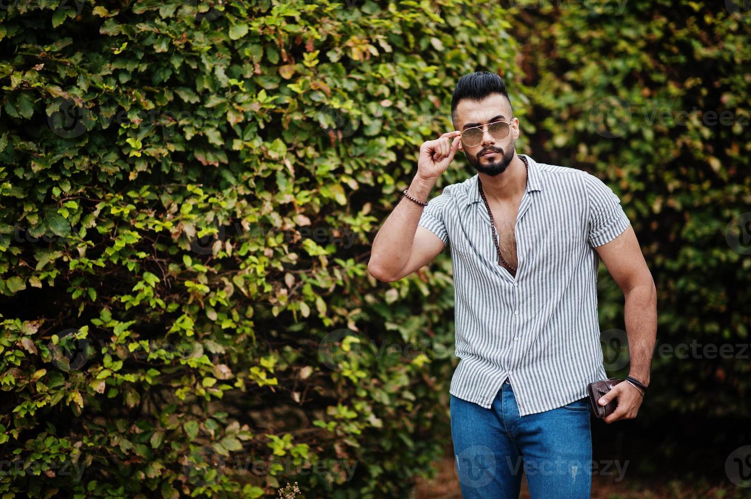 modieuze lange Arabische baard man slijtage op shirt, jeans en zonnebril gesteld op park tegen groen. foto