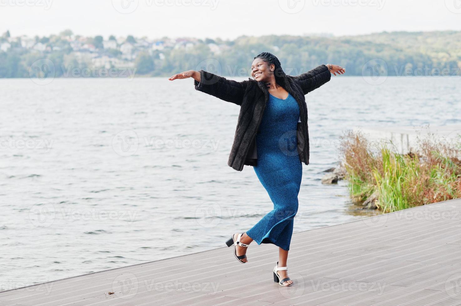 Afro-Amerikaanse donkere huid plus size model poseerde in een blauwe glanzende jurk en zwarte bontjas tegen de zeezijde. foto