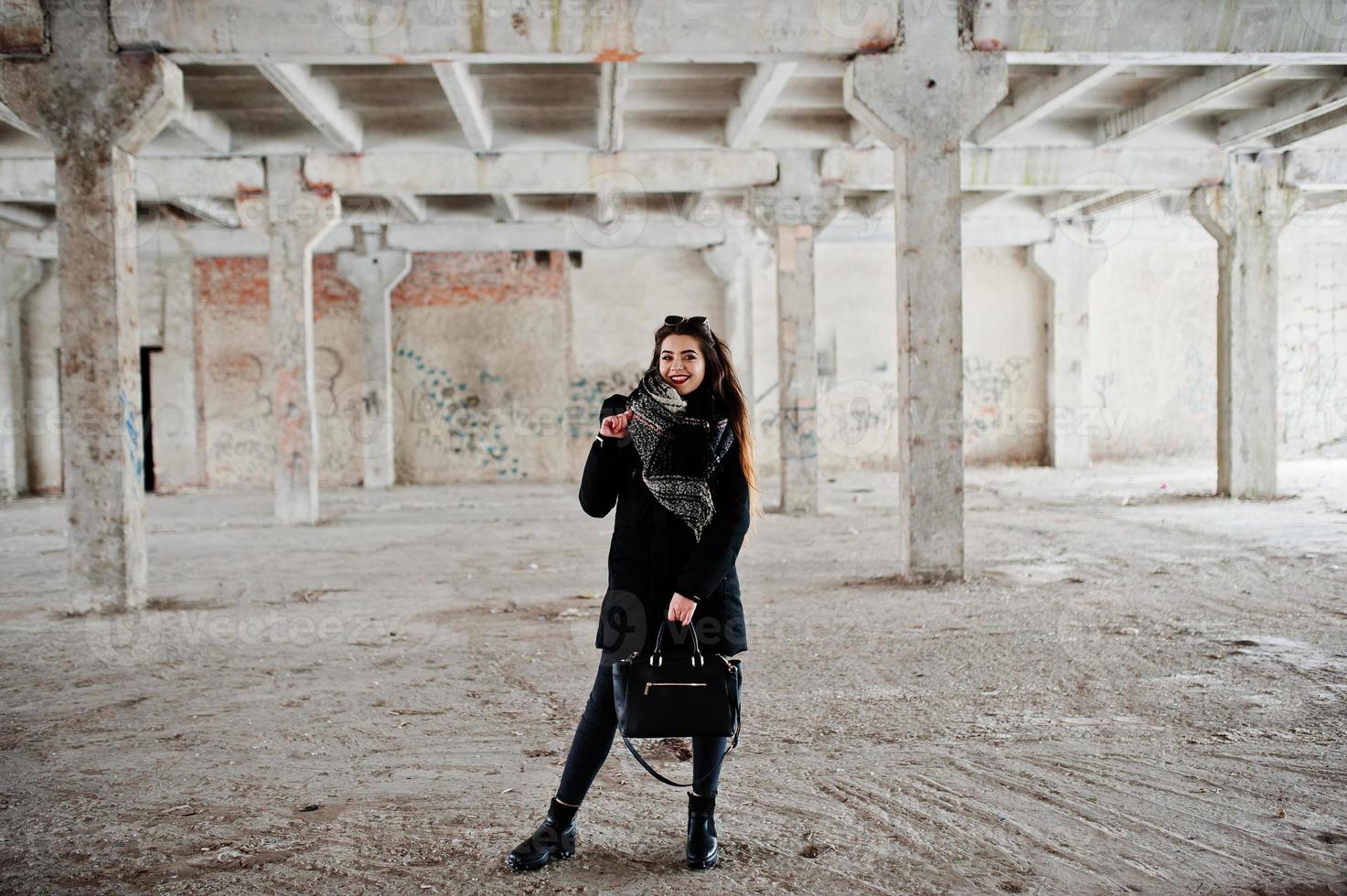 brunette stijlvolle casual meisje in sjaal en handtas tegen verlaten fabrieksplaats. foto