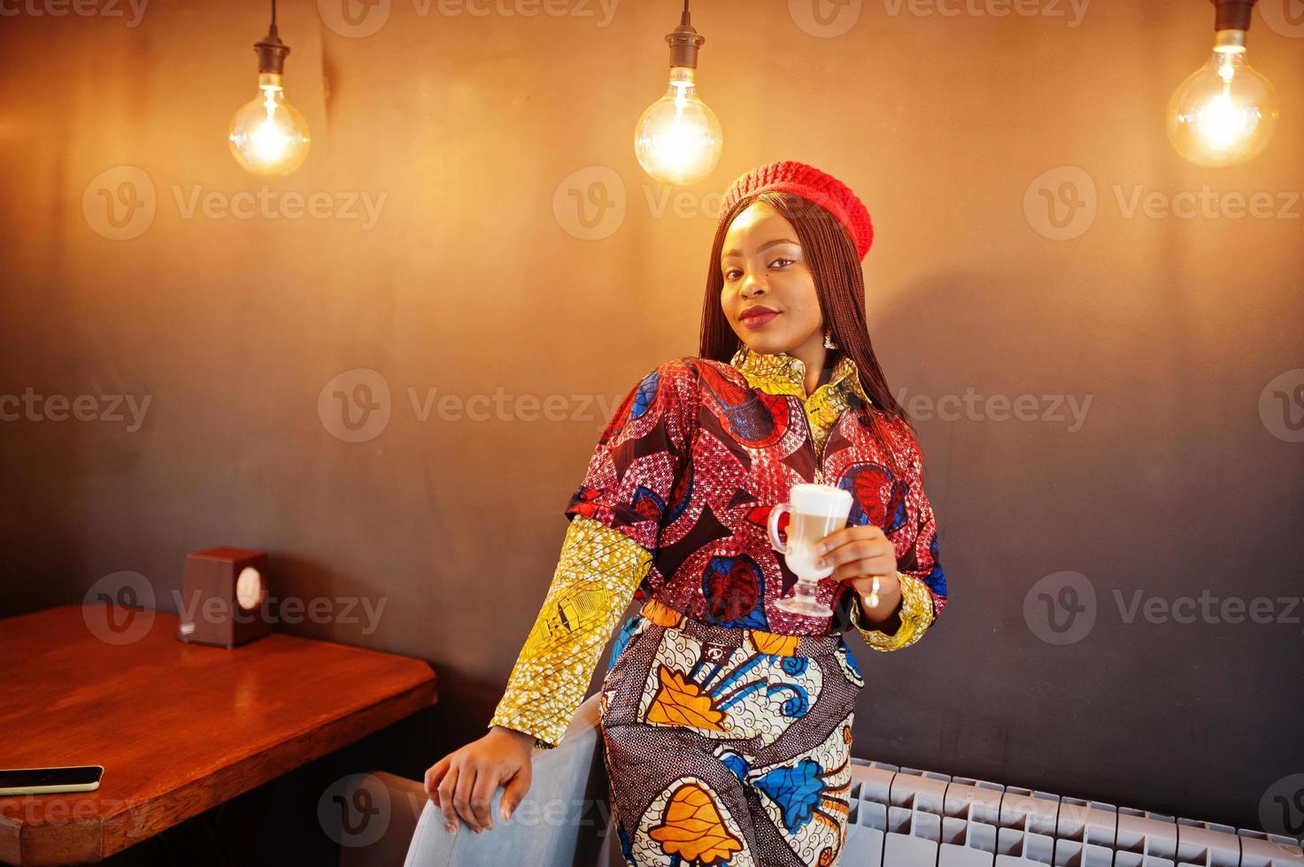 enthousiaste afro-amerikaanse vrouw in trendy gekleurde outfit met rode baret chillen in gezellig café met kop warme latte in handen. foto