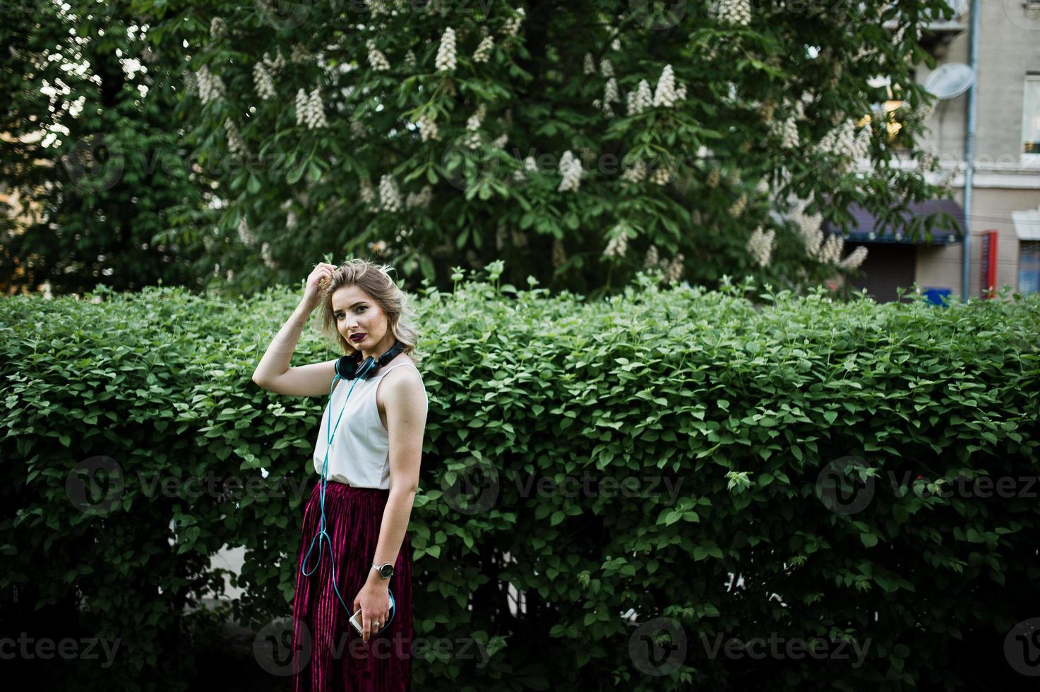 modieuze en mooie blonde model meisje in stijlvolle rood fluwelen velours rok, witte blouse, geposeerd in het park met telefoon en koptelefoon. foto