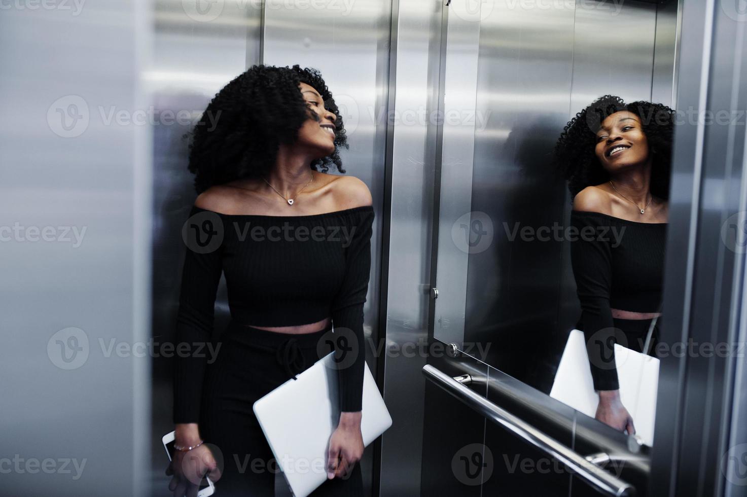 modieuze jonge mooie Afro-Amerikaanse zakenvrouw met afro kapsel dragen in elegant zwart, staan bij de lift met telefoon en laptop bij de hand. foto